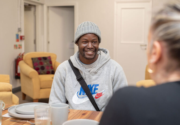 Gentle man sat in Tabor lounge, wearing grey hooded jacket and beany. Smiling towards staff member.