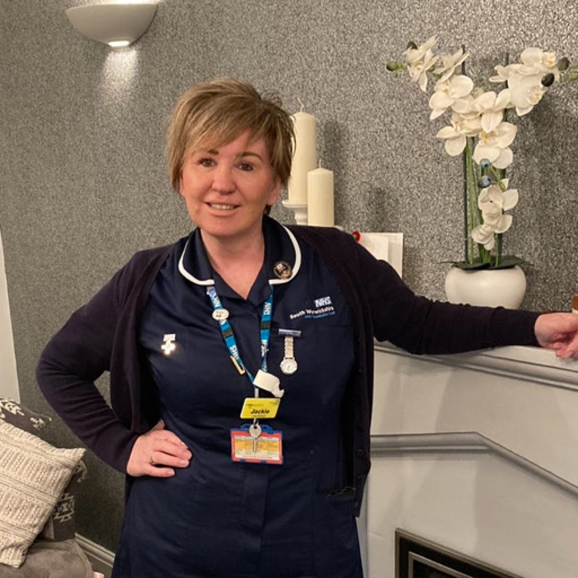 Lady in dark blue nurse uniform with short hair smiling ad leaning on a mantle piece