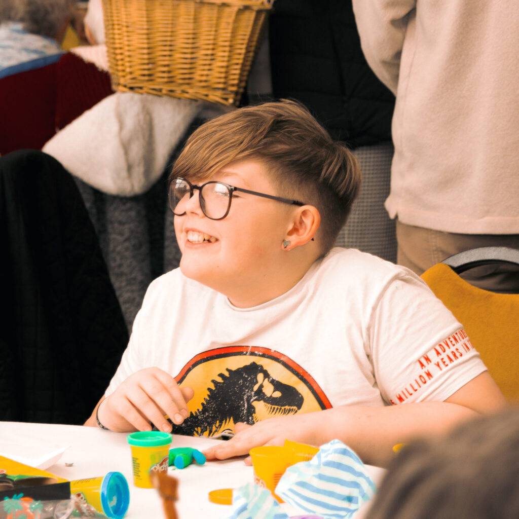 Young boy wearing glasses and a dinosaur t-shirt playing with playdough and smiling away from the camera