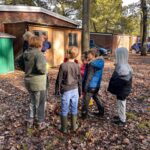 Autumnal picture of Children standing in a circle in the woods with leaves on the floor. Children dressed in welly's and coats, looking towards adults.