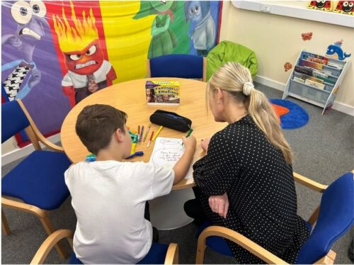 Child and support worker sat at round table. Child is drawing and adult is looking eagerly at their drawing